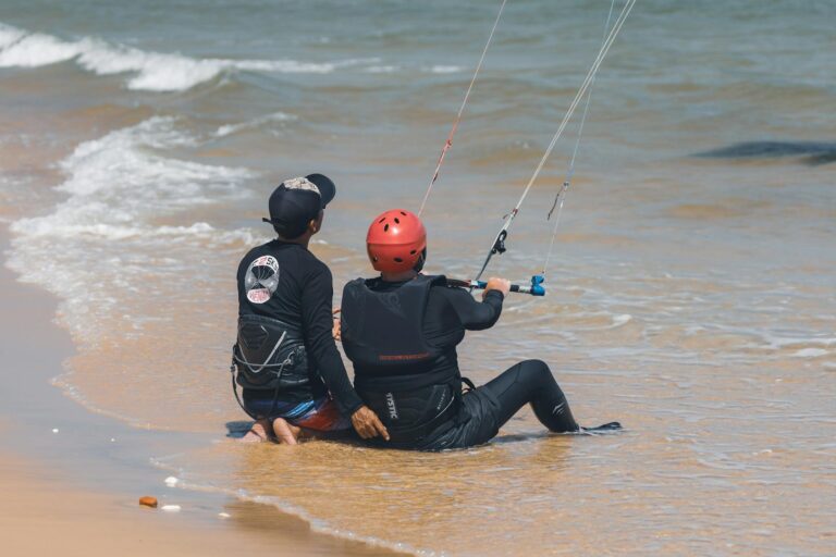 Two people in wetsuits receiving kitesurfing lessons on a sunny beach shoreline.